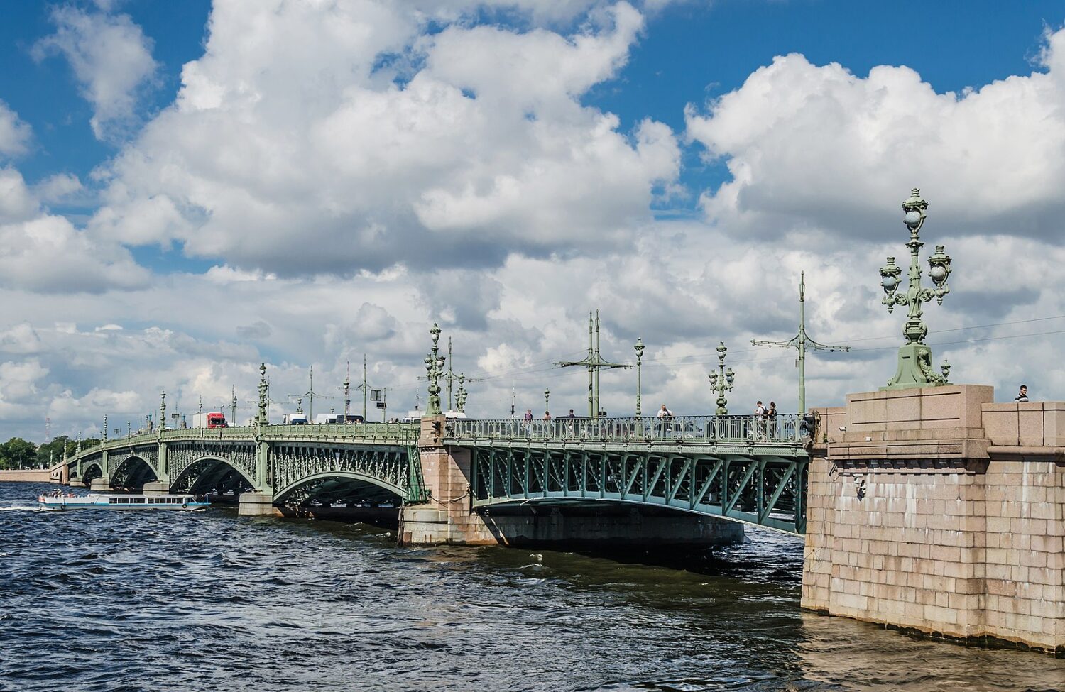 Un pont de métal vert avec quelques colonnes de pierres beiges s'étend au-dessus d'une eau bleu-gris agitée. Un ciel légèrement bleu, mais principalement ennuagé, se profile au-dessus du pont. 