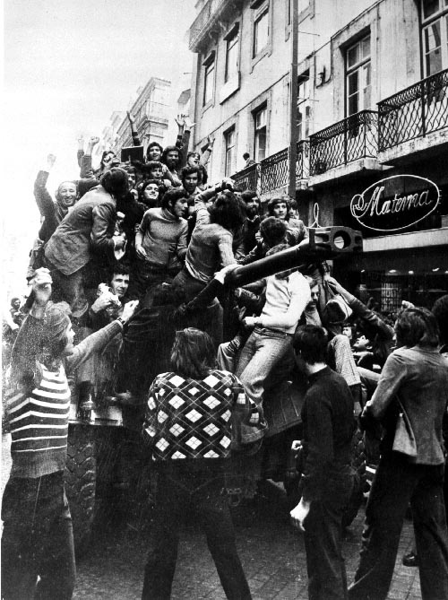 Une photo en noir et blanc montre une foule de jeunes hommes civils, principalement, monte sur un char d'assaut. Ils ont les bras en l'air en signe de victoire et semblent célébrer. Le tout se passe devant des édifices, qu'on voit à droite de la photo. Ils semblent être des appartements et un café ou une boutique.