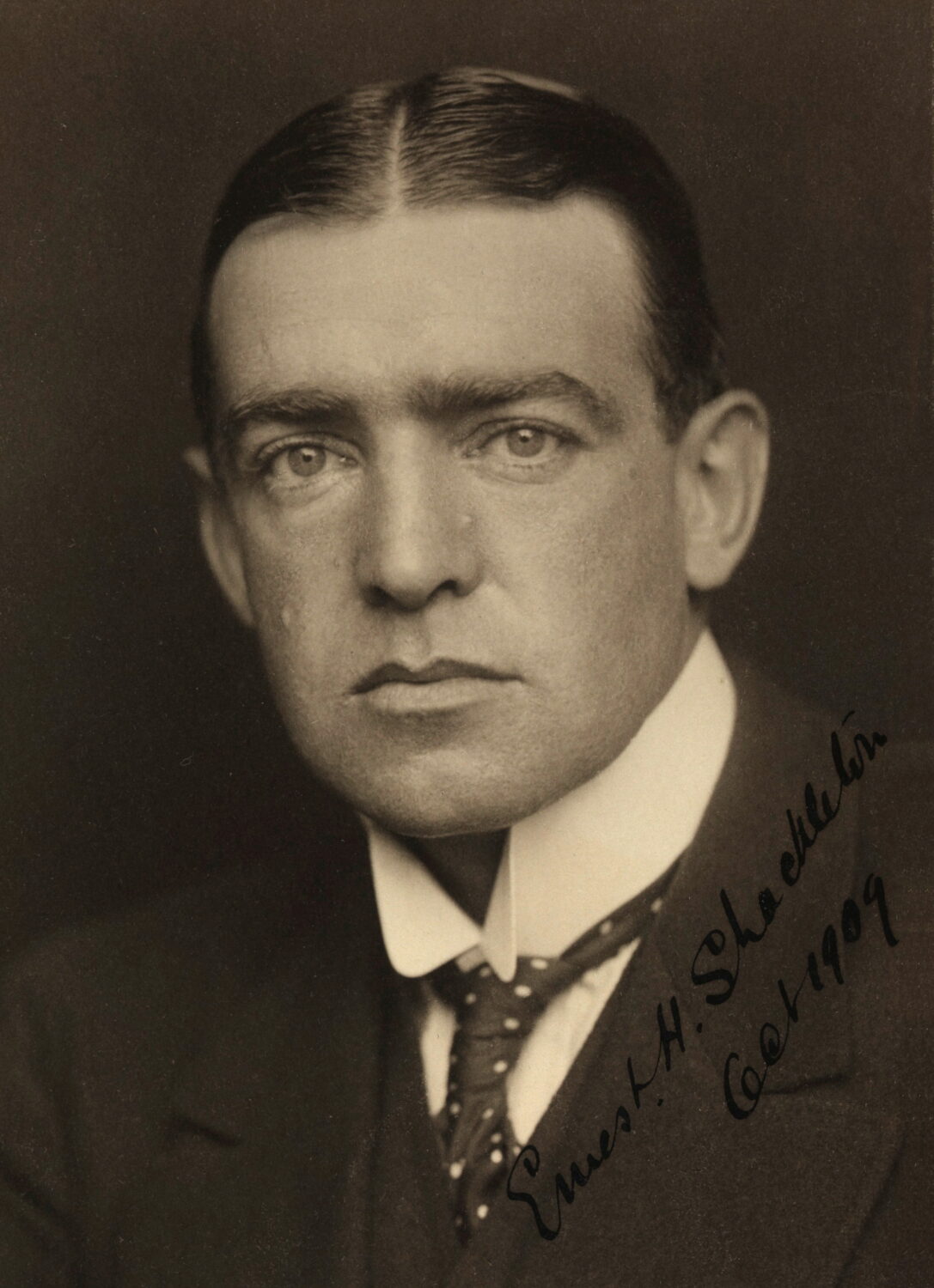 Une photo de Shackleton prise en sépia. Il regarde la caméra de face. Ses cheveux sont graissés et séparés par une ligne bien visible au centre de son crâne.