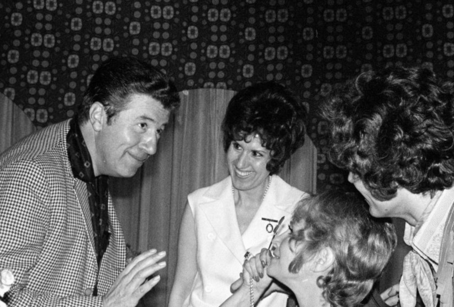 Une photo en noir et blanc montre un homme habillé d'un veston carreauté et d'une chemise foncée penché vers trois jeunes femmes avec qui il discute. 