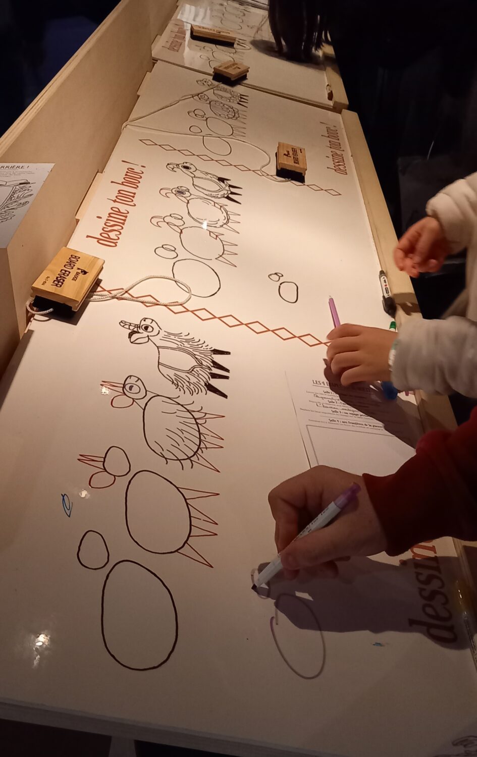 Une longue table avec des papiers blancs sur lequel sont dessinés des ronds qui se transforment en bouc, étape par étape de dessin expliquée, se trouve devant des enfants dont on ne voit que les mains qui dessinent lesdits boucs.
