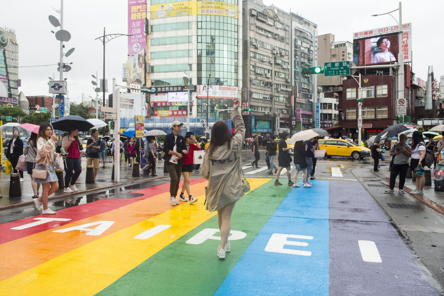 Un jeune femme dans un manteau beige marche sur une rue peinturée aux couleurs de l'arc-en-ciel sur laquelle Taipei est écrit en blanc. Elle fait dos à la caméra. D'autres passants, plusieurs avec des parapluies, marchent autour d'elle. En arrière-plan, de grands édifices vitrés et deux voitures jaunes. 