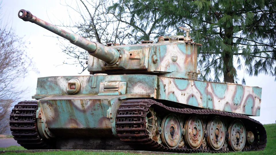 La photo montre un tank beige avec de grosses taches bleu-vert. Il fait face vers la gauche et est placé devant des arbres que l'on voit se détacher, en arrière-plan, d'un ciel gris.