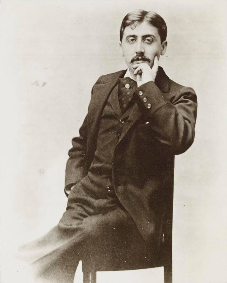 Une photo en noir et blanc montre Marcel Proust assis de côté sur une chaise. Son coude gauche est accoté au dossier de la chaise et sa tête repose sur ses doigts. Sa main droite est dans sa poche de pantalon. Proust porte un tailleur de couleur foncé avec un foulard foncé au motif picoté plus pâle. Il regarde fixement l'appareil, ses cheveux bien peignés et sa moustache aussi. 