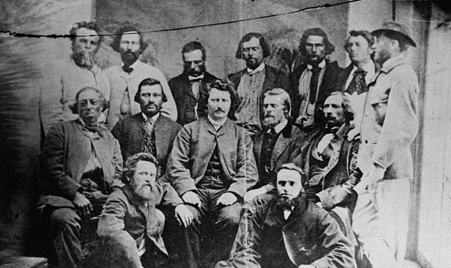 Un groupe de 14 hommes est photographié en noir et blanc. Ils portent tous un complet, certains en couleurs foncées d'autres en couleurs pâles. Certains regardent la caméra et d'autres non. Louis Riel, assis au milieu du groupe, regarde fixement la caméra. Les hommes sont disposés en trois rangées : la première est composée de 7 hommes debout, la seconde de 5 hommes assis sur un banc et la dernière de 2 hommes assis par terre.