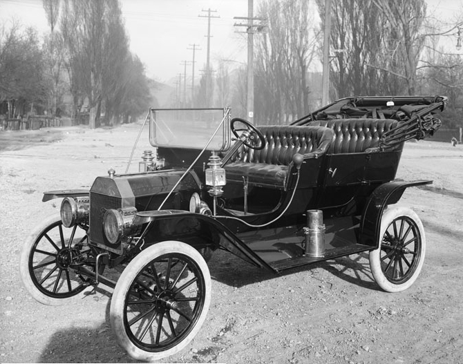 La photo en noir et blanc présente une voiture antique au milieu d'une route avec des arbres et des poteaux électriques de part et d'autre du chemin dégagé. La route est en terre batture. La voiture n'a pas de toit ou, du moins, a un toit en toile qui est replié à l'arrière. Elle n'a pas de portes non plus. Elle est d'une couleur foncée et présente deux sièges qui ressemblent à des banquettes, l'un derrière l'autre. Le volant pour le conducteur se trouve à gauche. Les quatre roues de la voiture sont assez minces et pâles.