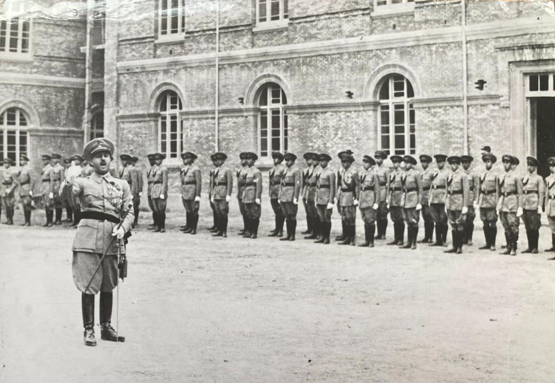 La photo en gris montre Franco en uniforme militaire devant un groupe de soldats. Franco est en plein milieu d'un discours, la bouche grande ouverte et la main droite légèrement levée, à la hauteur de son épaule. Les soldats se tiennent en ligne, le dos bien droit et les bras le long du corps. Ils font dos à un bâtiment imposant, composé de briques et ayant de nombreuses fenêtres en arche. On aperçoit la moitié d'une porte rectangulaire et ouverte tout à fait à droite de la photo. 