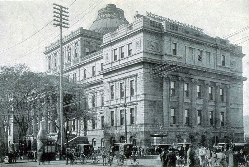 La photo en noir et blanc montre un édifice carré et imposant. On le voit de l'autre côté d'une rue, de biais, se concentrant sur un coin qui montre la façade et un côté du bâtiment. Un dôme se dresse au milieu de l'édifice. De nombreuses fenêtres se trouvent sur les deux façades, celles de côté encadrées par des colonnes cannelées de style romain. Devant la façade avant, un grand poteau électrique. Au pied de l'édifice, vis-à-vis le coin, se trouve une fontaine avec une statue sur le dessus. De nombreuses charrettes avec des chevaux attelés attendent au pied de l'édifice, de même que plusieurs piétons. 