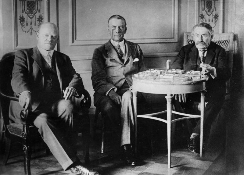 La photo en noir et blanc présente trois hommes en complets assis sur des chaises devant une petite table ronde remplie d'objets divers. Les trois hommes font face à la caméra. Celui du milieu porte un monocle et celui de droite a une moustache bien fournie.